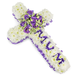 This solemn floral cross is composed with close attention to texture and messaging: a substantial foundation of small, white chrysanthemums forms a seamless, cushion-like surface that reads both pure and steady against the stark white backdrop. A ruffled, deep purple satin ribbon is woven around the entire perimeter, its satiny sheen and pleated edge lending a formal, respectful border. Down the vertical arm the letters "MUM" are carefully spelled by clusters of compact purple blooms-miniature lisianthus or spray chrysanthemums-each flower selected for colour consistency so the dedication reads clearly from a short distance. The central spray is a richly layered mini-bouquet of deep violet roses and lighter lavender lisianthus, their petals creating a gentle contrast of tones and a soft focal glow, complemented by variegated greens with light-edged leaves and a scattering of rounded green berries for tactile contrast. There is a subtle floral fragrance-cool and powdery-that feels consoling rather than overwhelming. This design balances simplicity and craftsmanship, appropriate for a funeral service at a local church near Stanmore tube or a memorial at Canons Park cemetery. Prepared with experienced hands and available for local delivery across Stanmore and neighbouring Harrow, this cross conveys dignity, sincere condolence, and a personal tribute to a beloved mother, executed thoughtfully by Flowers Stanmore with the neighbourhood's quiet care in mind.