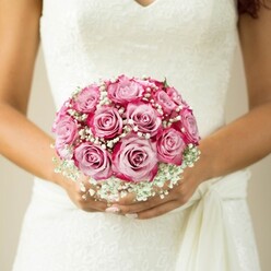 Bathed in gentle light, the artistry of a Stanmore bridal bouquet is revealed: a symphony of pink roses in full, radiant bloom, their petals fading seamlessly from blush heart to vivid fuchsia rim. Each rose, hand-selected for peak freshness from local markets near Stanmore Hill, is intertwined with sprays of airy white baby's breath, adding a dew-kissed, almost ethereal finish. The bouquet is quietly cradled by the bride-her hands warm and graceful, nails polished in the palest shimmer-reflecting the serenity of weddings held amid Stanmore's green parks or stately gardens. Her dress, a sleeveless vision of floral lace with a flowing sash, echoes time-honoured bridal fashion as seen throughout Stanmore's wedding venues. The neutral backdrop gently recedes, focusing every sense on this arrangement's tactile beauty and inviting the onlooker to imagine the gentle fragrance of fresh roses as they mark the start of a lifelong journey. Flowers Stanmore's meticulous craftsmanship is evident in every bloom, radiating hope, tradition, and a quiet joy perfect for wedding celebrations in north-west London.