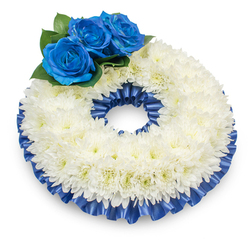 Captured from slightly above on a white background, this circular wreath presents a composed and dignified expression of sympathy, shaped by hundreds of delicate white chrysanthemums whose feathery petals form a full, textured ring. The chrysanthemums' soft surface is subtly warmed by faint creamy centers while remaining overwhelmingly serene. At the upper-left portion of the ring, three electric blue roses rise like a focused point of remembrance: richly saturated, velvety petals tightly spiralled to suggest devotion and solemn beauty. Dark green leaves cradle the roses, giving visual weight and a natural contrast to the luminous whites and deep blues. Encircling the wreath is a pleated cobalt satin ribbon, its ruffles catching light and shadow to define both the outer and inner edges with a polished finish. The composition reads as both traditional and personal - appropriate for funeral chapels, graveside tribute displays, or quiet services in local venues around Stanmore and Harrow. I prepare pieces like this with attention to texture, scent, and proportion; the mild fragrance of chrysanthemums and the gentle musk of the roses lend the wreath a comforting presence without overwhelming the space. Flowers Stanmore can prepare and deliver similar tributes across Stanmore, to addresses near Stanmore Station or Canons Park, ensuring a respectful, timely offering for those intimate moments of farewell and remembrance.