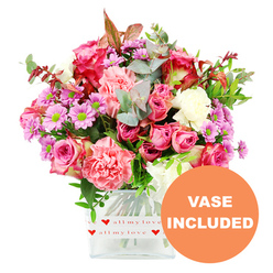 This image shows a generous, hand-crafted bouquet displayed in a square clear glass vase, photographed head-on against a clean white background that echoes the tidy elegance of a Stanmore living room. The arrangement is a celebration of pinks: large, velvety roses open in layers of deep fuchsia and magenta alongside softer blush and pastel shades, giving the bouquet a romantic, storytelling feel. Between them, plump, ruffled carnations in coral-pink and peachy tones form cushions of texture, like soft fabric folded around the roses. Sprinkled throughout are delicate, daisy-style chrysanthemums in lavender and light purple, their vivid lime-green centres providing bright pinpoints of contrast that lift the entire colour scheme. Creamy white blooms lie tucked between the stronger hues, adding a sense of air and spaciousness. The flowers are held together by plentiful greenery: smooth, rounded eucalyptus leaves in soft blue-green, richer dark green foliage, and a few small sprays of reddish-brown leaves that give the design a slightly seasonal, intimate note. The clear glass vase, with its neat, contemporary lines, is wrapped at the base with a white band decorated in a repeat pattern of red hearts and the phrase all my love in flowing red script, making the container itself part of the message. In the bottom right corner, an orange circular emblem with bold white capital letters reads VASE INCLUDED, drawing attention to the ready-to-display nature of the design. The overall impression is of a warm, caring bouquet you might have delivered to a home off Stanmore Hill, an apartment near the station, or a family house close to Canons Park, to celebrate a birthday, anniversary, or simple everyday affection.