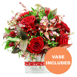 This image shows a sumptuous, love-themed arrangement placed against a pure white backdrop, its reds and greens glowing as though lit by soft daylight. At the center, several large red roses unfurl in different stages of bloom, their velvety petals ranging from tightly furled spirals to fully open, layered rosettes, conveying both drama and tenderness. Echoing their passionate hue, deep crimson accent flowers, likely alstroemeria or a similar variety, nestle between the roses, their slender petals and intricate markings creating movement and subtle pattern. Threaded between these stronger elements are delicate sprays of tiny pale pink and white waxflower-like blossoms, with minuscule cups and buds that resemble a dusting of floral lace, lightening and softening the bouquet. Enveloping the blooms is an array of lush greenery, from broad, smooth leaves to finer, more textured sprigs, which provide fresh contrast and structure. Striking burgundy and reddish-brown leaves, some gently curved and finely veined, extend outward, introducing a note of refined drama that feels well suited to a stylish home near Stanmore station or along The Broadway. The composition forms a full, rounded dome, thoughtfully constructed to appear generous from every angle, reflecting the practiced hand of an experienced florist. All the stems are arranged within a sleek, clear rectangular glass vase, where the straight edges offer a contemporary counterpoint to the natural forms of the flowers. A red ribbon adorned with repeating white heart outlines encircles the upper portion of the vase, quietly reinforcing the romantic message. In the bottom right corner of the image, a circular orange overlay displays the phrase "VASE INCLUDED" in bold white lettering, reassuring anyone ordering this bouquet for delivery in Stanmore that the gift comes complete, ready to place on a dining table, bedside, or office desk.