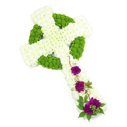 In this image, a large Celtic cross floral arrangement is shown laid flat against a clean white studio background, each part of the design carefully defined. The main structure of the cross is created from countless small white chrysanthemums, densely packed together so that no gaps show, forming a continuous, cushion-like surface that follows the vertical stem and horizontal arms. At the centre, the classic halo of the Celtic cross stands out as a strong ring of lime-green button chrysanthemums, their small, rounded heads forming a vivid band of colour. These green blooms carry on into certain segments of the arms and inner shapes, hinting at interwoven knot patterns often chosen by families in Stanmore with Celtic roots or an appreciation for traditional symbolism. The contrast between the fresh green and pristine white suggests balance and renewal, much like the calm greens of Canons Park after a gentle rain. On the lower portion of the vertical arm, three distinct clusters of deep purple carnations bring a heartfelt, emotional note. Their ruffled petals are rich and plush, providing a deep, comforting colour that stands out yet feels harmonious with the rest of the design. Intermingled with these carnations are fine sprigs of dark green foliage and a few slender, twisting stems that trail gracefully across the chrysanthemums, giving the arrangement movement and a natural, garden-like touch. Soft, even lighting allows every petal and leaf to be seen clearly, reflecting a tribute freshly prepared by Flowers Stanmore for delivery to a local church, chapel, or family home. The cross conveys a powerful blend of remembrance, faith, and peace, suited to funerals, memorials, or celebrations of life in the Stanmore area.