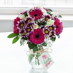 A lush, joy-filled bouquet of mixed flowers rests in a squat, textured clear glass vase, framed by a luminous white background that suggests soft daylight streaming through a window in a quiet Stanmore side street. At the heart of the arrangement, bold gerbera daisies in saturated fuchsia and magenta create an immediate sense of celebration, their dark centres adding depth and drama. These statement blooms are encircled by generous clusters of daisy-like chrysanthemums, each petal tipped in velvety purple and fading gracefully to white as it approaches a bright yellow centre, echoing the cheerful feel of summer walks through Bentley Priory Nature Reserve. Scattered between the stronger hues, creamy white roses appear in various stages of opening, from tight buds to fuller blooms, lending an elegant, timeless note and a suggestion of gentle fragrance. Wisps of airy white baby's breath weave among the larger flowers, adding a gossamer lightness, while rich green foliage fills out the base and sides, giving the bouquet a full, balanced silhouette and a natural garden character. The clear glass of the vase reveals the neatly trimmed stems and a hint of water, reinforcing the impression of freshness and careful florist craftsmanship. Around the neck of the vase, a length of rustic twine is tied into a simple bow, from which hangs a small, light-toned gift tag adorned with a soft pink floral emblem and delicate script that implies a thoughtful message for the recipient. Illuminated by gentle, diffused light, each petal catches a subtle glow, making the colours appear vivid yet harmonious. The composition evokes the feeling of a lovingly prepared floral gift, the kind that might be delivered across Stanmore to mark a birthday, say thank you, or bring comfort on a quiet evening at home.