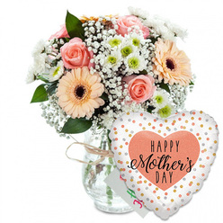 This photograph presents a tender Mother's Day gift set, thoughtfully composed and shown against a pristine white backdrop that echoes the calm, open feel of Stanmore's residential streets. Dominating the left side of the frame, a lush mixed bouquet rises from a clear glass vase, the stems visible through the water like a neat bundle gathered by an experienced florist from a studio near Stanmore Broadway. The vase is finished with a simple loop of rustic twine at its neck, adding a quiet, handcrafted note that suggests time and care have gone into this gift. The bouquet itself is rounded and bountiful, packed with soft, romantic colours. Fully opened peach and blush-pink roses showcase spirals of velvety petals, each bloom inviting you to picture the soft touch of the flower and the comfort of giving it to someone dear. Around them, bright peach gerberas with deep, dark centres bring in a more playful element, their wide, symmetrical faces reminiscent of the cheerful energy of a family gathering. Between the larger blooms, a light mist of white baby's breath softens the lines of the arrangement, creating a dreamy, cloud-like effect that makes the bouquet feel light and airy. Small white chrysanthemums with pale green hearts nestle in the gaps, adding texture and giving the arrangement resilience and shape, much like the layered greenery you might find on a walk through Bentley Priory Nature Reserve. Deep, glossy foliage surrounds and supports the flowers, enriching the palette and suggesting freshly conditioned stems. In the foreground to the right, a large heart-shaped foil balloon adds unmistakable celebration. Its white surface is sprinkled with metallic gold and silver dots, scattered like confetti tossed into the air. At the centre, a warm peach heart frames the words "HAPPY Mother's DAY" in bold black type, the word "Mother's" written in flowing, calligraphic script that brings intimacy to the message. A small white card tucked behind the vase and balloon hints at a personal note. Together, the bouquet, balloon, and card create a complete visual story of appreciation, ideal for a Mother's Day delivery to a home anywhere in Stanmore.