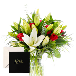 This bouquet is a portrait of sophistication and emotion, thoughtfully composed to evoke admiration and comfort in equal measure. The white lilies stand regal at the heart, their large trumpet shapes unfurling among abundant green spears and curls of foliage. Red roses cluster throughout with a fervent, almost cinematic romance, their fragrance mingling with the lilies in a dewy, hypnotic whisper. The abundance of unopened buds promises beauty still to come, a sense of hopeful ongoing celebration. The natural arrangement is caught in bright, clear light, each leaf and petal outlined with freshness as if sketched by a careful hand. A graceful card marked Hero completes the ensemble, making it an ideal expression of thanks or remembrance for someone in Stanmore whose spirit inspires. The bouquet sits beside a quiet coffee table on The Broadway, ready for same-day delivery across Stanmore and the surrounding Harrow area. Its presence speaks of respect, gratitude, and quiet strength, a refined gesture suitable for anniversaries, congratulations, or a heartfelt note to a friend who has risen to a challenge. Set in a slender glass vase, the composition balances form and texture, inviting the eye to follow the curves of the stems and the glow of the petals from Stanmore Hill to Canons Park.