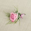 A single blush pink rose boutonniere is displayed from a slightly elevated angle on a softly textured, light beige linen background, the neutral fabric reminiscent of a sunlit table in a Stanmore townhouse near Belmont Circle. The rose is semi-open, its velvety petals spiralling gently outward from a tighter centre, where the colour is a tender pastel pink that gradually deepens to a more vibrant, romantic hue along the slightly curled outer edges. Around the base of this central bloom, tiny white baby's breath blossoms cluster like a delicate cloud, their miniature, pearl-like flowers adding lightness and contrast without overpowering the rose. Wispy, emerald green strands of grass-like foliage fan out gracefully from behind, creating a sense of movement and an airy frame, while a single broader, darker green leaf is folded neatly to cradle the stems, lending structure and a tailored finish. The short stems are carefully wrapped in a lustrous satin ribbon, blending soft lavender-pink with a deeper magenta-purple tone, tied in a secure, simple knot that mirrors the refined style often chosen for weddings at local Stanmore venues or school proms nearby. Soft, even lighting casts the slightest of shadows, emphasising the texture of the petals, the woven linen, and the smooth sheen of the ribbon. This intimate close-up captures the calm romance of a handcrafted boutonniere, ready to sit on a groom's lapel, a groomsman's jacket, or a young guest's suit as they step out onto the quiet streets around Stanmore Hill for an evening of celebration and photographs in the gentle North London light.