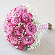 A softly lit image shows a hand-tied bouquet of roses and baby's breath resting diagonally on a pale, linen-like cloth, reminiscent of a serene morning in Stanmore before guests arrive for a celebration. At the heart of the bouquet, around a dozen roses form a rounded, almost domed centre, their petals blending from cooled lavender and dusty mauve tones at the core to vivid fuchsia and bright magenta at the edges. The variation in colour creates an ombré effect, each bloom appearing almost three-dimensional as the petals curl and twist in graceful whorls. The roses are closely packed, giving the arrangement a lush, full appearance. Wrapped around them is a generous cloud of baby's breath, the countless tiny white blossoms of gypsophila filling the space between the larger flowers like a delicate veil. This airy halo softens the intensity of the pinks and purples, lending the bouquet a dreamlike quality. Dark green leaves are mostly hidden beneath the blooms, but subtle glimpses of foliage peek through, providing quiet contrast and a reminder of the freshness of the flowers. The stems are carefully aligned and wrapped in a lilac satin ribbon, its smooth surface gently reflecting the light. A spiral of small white pearls winds along the length of the ribbon, giving a distinctly bridal and formal feel. The background fabric, a light beige or off-white with a soft texture, adds warmth to the composition without drawing attention away from the flowers. This bouquet would suit a wedding at a Stanmore synagogue, a civil ceremony at a local registry office, or an intimate anniversary celebration at home, evoking feelings of tenderness, devotion, and refined taste.
