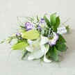 An elongated hand-tied bouquet is laid diagonally across a softly textured, light beige fabric, its gentle neutral tone allowing the flowers to glow as the clear focal point. At the heart of the design, several large white Oriental lilies open fully, their silky petals curling back to reveal golden-yellow stamens dusted with pollen and fine reddish speckles that add character to each bloom. Around them, smooth chartreuse-green buds sit poised to open, suggesting movement and life still to come. Threaded between the lilies are ruffled Lisianthus blossoms in white kissed with vivid purple edges, some shaded from lavender to deep violet in their centres, giving the arrangement a modern, painterly feel. A scattering of fine baby's breath creates a faint, cloud-like haze, softening the edges of the bouquet and adding a romantic whisper. Rich, layered greenery in varied shapes and finishes-broad, glossy leaves, slender blades, and feathery fronds-fans out to frame the blooms, adding depth and texture. The stems are neatly aligned and cinched with a crisp white satin ribbon, delicately embellished with tiny pearl-like beads along the narrower right-hand end, echoing the elegance of a bridal gown. Soft, even lighting highlights the clean whites, fresh greens, and jewel-like purples without harsh shadow, creating a calm, timeless mood. The bouquet feels perfect for a Stanmore bride walking from home towards Bentley Priory or a special anniversary dinner at a favourite local restaurant, its fragrance an imagined blend of lily sweetness and green freshness. The scene conveys careful craftsmanship and quiet celebration, like something lovingly prepared by a trusted florist who understands local weddings and heartfelt gifts in Stanmore.