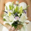 This Stanmore bridal bouquet is a sensory ode to love and new beginnings, composed with a poet's eye for colour and a florist's sense of harmony. White lilies-broad, soft, and radiant-unfurl at the centre, releasing their gentle perfume and capturing the serene character found in Stanmore's gardens and green spaces. Bunches of lisianthus curve around them, the white petals ruffled and touched with lavender as though brushed by twilight. Deep purple accents from veronica flashes create spark while fluffy mistlike tufts of baby's breath drift through the formation, invoking airy spring mornings. Lush greenery spills from the bouquet's edges-broad leaves, unopened buds, and feathery asparagus fern-all arranged to evoke movement and freshness, reminiscent of nature walks around Canons Park or in Bentley Priory Nature Reserve. The bride's manicured hands, adorned with light pastel polish, cradle the bouquet, while the white of her strapless dress glows softly behind, bringing attention to every thoughtful floral detail. Crafted with care by Flowers Stanmore and inspired by local landscapes, this bouquet is a radiant testament to celebration and enduring love, designed for memorable Stanmore weddings.