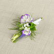 A small composition with a softly romantic voice, this boutonniere pairs two white lisianthus blooms rimmed in pale violet with airy sprays of baby's breath to create a feeling of restrained celebration. The lisianthus present as tactile, slightly ruffled petals - cool and satiny to the touch - while the tiny white filler flowers provide a mist-like halo that lifts the arrangement and suggests delicate movement. Rising beneath them are narrow stems of grape hyacinth, their blue-violet spikes adding pinpoint contrast and a vertical rhythm that balances the rounded blooms above. Slender buds and fresh pale shoots extend like quiet punctuation marks, and wisps of fern introduce a woodland softness, their feathery fronds catching glints of light and tempering the colour palette with soft green. The bunch is fastened with a fine white satin ribbon and set against a neutral linen backdrop whose woven threads create a gentle textural counterpoint to the smooth petals. Prepared with care in our Stanmore studio at Flowers Stanmore, this boutonniere is meant to be experienced up close - the velvety petals, the faint floral perfume, the smooth satin against a lapel - and is ideally suited for weddings, anniversaries, or a formal portrait taken near Stanmore tube station or the green paths of Canons Park. It reads as thoughtful rather than flashy, an understated flourish that holds attention in quiet, personal moments and complements both contemporary tailoring and classic suits.