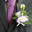 Nestled against the deep charcoal cloth of a well-cut suit, this boutonnière feels like a careful promise folded into fabric. The focal flower is a white lisianthus, its petals buttery and soft, each rimmed with a luminous purple that gives the bloom an unexpected, refined edge; nearby a white dianthus with energetic purple ruffles adds a touch of cheer and lively texture, its serrated petals catching tiny highlights. Behind them, three slender lisianthus buds stand ready to open, a quiet suggestion of beginnings, while a single stem of dark purple veronica lends a vertical accent and a touch of vintage charm. Lush dark-green foliage and loops of ornamental grass form a natural frame, their blades glossy and slightly translucent where the light passes through. Scattered among the stems, tiny clusters of gypsophila read like confetti - soft, cloudlike, and breathing light into the composition. A neat white ribbon ties the stems with restraint and refinement, echoing the boutonnière's palette of white and violet. When pinned beside a magenta silk tie, the arrangement becomes both an anchor and a flourish, suited for a groom or honoured guest at a Stanmore ceremony, whether near Canons Park or along Stanmore Broadway. As Flowers Stanmore's florists would tell you, the piece is crafted for endurance as well as beauty: it offers a faint, fresh scent and a steady presence through long days of celebration, a tiny living emblem of care and tasteful confidence.