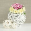 A serene, close-up scene shows a romantic rose centrepiece poised on a pristine white surface, the sort of arrangement that could brighten a Stanmore home near The Broadway or enhance a wedding table at a nearby country club. At the heart of the image lies a dome of fresh roses, thoughtfully combined in two complementary shades. The upper section of the dome is filled with pastel pink and soft lavender-pink blooms, their petals unfolding in gentle spirals that suggest softness and intimacy. Around the lower edge, a ring of creamy ivory and buttery pale yellow roses adds warmth and a subtle glow, helping the colours blend seamlessly. The roses vary in openness, from tight buds to generously open blooms, giving the arrangement a natural, just-gathered feel. Interlaced between them are delicate sprigs of white baby's breath, whose tiny blossoms create a misty, almost cloud-like texture that lightens the entire composition. Supporting the flowers is a distinctive spherical vase crafted from white-painted wicker or fine branches, woven together into an open, airy globe. Its lattice structure offers glimpses of the inner container and contributes a handcrafted, rustic-meets-modern charm. To the left of the vase, two decorative spheres made from pale natural wood strips rest on the same white tabletop, their open-weave pattern echoing the design of the main vessel and adding visual balance. The background is a soft beige or light taupe wall, subtly textured like linen or gentle wallpaper, which allows the soft hues of the roses and the crisp whites to stand out. The overall mood is one of understated romance and careful styling, ideal for illustrating Stanmore wedding ideas, intimate celebrations, or inspiration for refined floral décor at home.