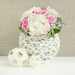 The image presents a beautifully composed bridal-style bouquet set against a calm, neutral background, the sort of arrangement that would look perfectly at home in a light-filled Stanmore venue near Canons Park. The flowers are gathered into a rounded, compact shape, suggesting careful, professional design. At its heart, creamy white peonies bloom in full glory, their petals layered thickly like folded silk, creating a sense of luxurious softness. Nestled among them, vibrant fuchsia-pink roses unfurl in tight spirals, contributing depth and a splash of vivid, joyful colour that immediately draws the eye. Fine sprays of baby's breath weave through the larger blooms, their tiny white flowers forming a delicate haze that lends the bouquet a romantic, almost ethereal aura. Around the base, broad, rich green leaves curve gently, adding structure and a fresh, botanical contrast to the pink and cream tones. The bouquet rises from a distinctive, spherical white vase, meticulously crafted from interwoven thin branches or rattan in an open lattice pattern that feels both contemporary and organic. Through its gaps, you can see shadows and hints of the stems, which enhances the sense of lightness and dimension. On the smooth white tabletop in front of the vase sit two decorative woven orbs, pale and airy, mirroring the vase's texture; one is slightly larger than the other, creating a gentle visual rhythm. The background is a softly textured beige, similar to fine linen or a carefully painted Stanmore home interior, allowing the flowers and woven elements to take centre stage. Illuminated by bright, even lighting, the entire scene radiates calm celebration and refined beauty, ideal for weddings, anniversaries, or special gatherings in the Stanmore area.