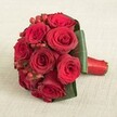 In the centre of this composition, a lush, compact bouquet of about ten large red roses draws the gaze with its vibrant, velvety textures and saturated crimson hues-craftsmanship that's typical of Flowers Stanmore's floral design. The roses are in full, elegant bloom, their delicate petals radiating shades of ruby and carmine beneath soft, diffused lighting that gently illuminates the arrangement. Bright red hypericum berries peek through the sea of petals, their round forms and glossy finish providing a playful contrast and festive accent, reminiscent of cheerful celebrations across Stanmore. Encircling the blooms, several broad deep green leaves are expertly curved to frame and support the flowers, mirroring the artful greenery found in Stanmore's open reserves. The stems are skillfully gathered and bound with a rich red satin ribbon, its sheen and snug finish offering both comfort and a polished appearance for carrying. Resting atop a neutral, textured linen fabric, the bouquet achieves a sense of timeless celebration, making it the perfect choice for weddings, anniversaries, or romantic occasions celebrated throughout the Stanmore area.