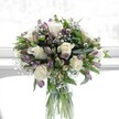 A softly lit floral arrangement stands in a clear cylindrical glass vase, placed against a bright, gently blurred background that suggests a sunlit window in a calm Stanmore home. At the heart of the bouquet, creamy white roses are fully open, their velvety petals spiralling outwards in perfect, romantic whorls that catch the light with a subtle sheen. Around them, clusters of lavender and muted purple alstroemeria, some still in tapered buds and others just beginning to unfurl, weave through the design with slender, speckled petals and delicate veining that deepens towards the throat of each bloom. Fine, cloud-like sprigs of white gypsophila, or baby's breath, fill the gaps between the larger flowers, giving the whole arrangement an airy, misty halo. Rich green foliage, a mix of broad glossy leaves and more slender stems, anchors the palette, framing the whites and purples and lending the composition a sense of freshness and movement, like a garden breeze drifting through Canons Park on an early spring morning. Through the transparent vase, the long green stems are clearly visible, freshly cut, criss-crossing in clean lines through the water and hinting at the careful craftsmanship behind the piece. The overall shape is full and rounded, designed with a florist's practiced eye so that every flower has space to breathe, yet the bouquet feels luxuriously abundant. The mood is serene, hopeful, and gently romantic, ideal for marking a new beginning, offering comfort, or celebrating a quiet milestone for someone in Stanmore or the nearby residential streets off The Broadway, where an elegant, restful arrangement like this can transform a room with understated grace.