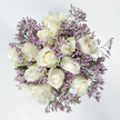 Against a clean, bright white backdrop, this overhead view reveals a softly rounded bouquet of white roses wrapped in a mist of lavender limonium, the sort of arrangement chosen to convey both purity and gentle remembrance. Approximately a dozen roses form the main structure, each bloom slightly different in its opening, which brings a natural, unforced rhythm to the design. Some of the roses remain closed and compact, with sleek, velvety petals that feel cool beneath the fingers. Others loosen into graceful spirals, their petals unfolding to reveal creamy white tones that deepen to subtle ivory, and in the very centre, a whisper of pale yellow warmth. A few of the roses have opened fully, forming delicate rosettes that seem to hover above the surrounding filler. Around and between them flows an airy border of limonium, its tiny lavender-purple flowers gathered densely along fine, branching stems. This filler forms a feather-light halo, adding both volume and an almost cloud-like softness to the bouquet, while providing a gentle colour contrast that never overwhelms the roses. Small glimpses of fresh green stems and discreet foliage appear here and there, adding hints of life and grounding the composition, yet the overall palette remains serenely limited to white, lavender, and soft green. The bouquet's mood is peaceful and reflective, much like the quiet residential avenues and green pathways that wind through Stanmore. Whether placed by a bedside, offered at a funeral service, or delivered to a family home near Stanmore Broadway, this arrangement offers a quiet embrace - a floral expression of sympathy, respect, and enduring love, conveyed through refined colour and tender, thoughtful craftsmanship.