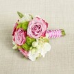 A small, hand-tied bouquet rests horizontally on a light tan, textured linen-style cloth, its colours glowing softly in gentle, even light. At the heart of the arrangement, three lush pink roses open towards the viewer, their petals spiralling layer by layer from tightly furled centres to wider outer petals. The hue shifts from a delicate blush pink in the middle to deeper, almost magenta-pink edging, creating a romantic ombre effect that feels both classic and modern. Nestled between the roses are rounded tufts of vivid lime green button chrysanthemums, their tiny, tightly packed florets forming cheerful, pom-pom shapes that add playful texture and a fresh, garden-picked feel. Threaded throughout the design are creamy white and soft ivory filler blooms, likely lisianthus or freesia, their silky petals and gentle ruffles lending lightness and a hint of sweet fragrance. Tiny star-like white blossoms with golden-yellow centres sparkle between the larger flowers like scattered confetti, enhancing the intricate detail. The stems are neatly trimmed and gathered into a compact handle on the right side, wrapped in a bright fuchsia satin ribbon that catches the light with a subtle sheen. Over the ribbon, a fine strand of white faux pearls is wound diagonally in a graceful spiral, suggesting bridal jewellery and adding a touch of refined glamour. The neutral fabric beneath, reminiscent of the table linens often seen at intimate Stanmore wedding venues near Bentley Priory or along Marsh Lane, keeps the focus firmly on the bouquet's pink, green, and white palette. The overall mood is one of fresh romance and understated celebration, perfect for a bridesmaid's posy, small bridal bouquet, or delicate centrepiece for an engagement or baby shower in Stanmore.