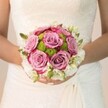 This scene captures a moment of quiet anticipation before a Stanmore wedding begins. In the foreground, the bride's gentle hands, polished in delicate pink and sparkling at the ring finger, support a stunning round bouquet, its blooms glowing in the filtered light often seen near Stanmore Hill or Bernays Park. At the bouquet's heart rest plush roses, their petals opening from soft lavender and dusty mauve at the center to deepening fuchsia at the rim, each flower imbued with subtle movement and depth. Encircling these, lively button chrysanthemums in lime-green mimic the vibrant lawns that characterise Stanmore's suburban gardens in early summer, bringing a touch of brightness and cheerful energy. A delicate outer layer of white blossoms and clusters-perhaps tiny freesia or spray roses-together with scattered green buds, finishes the ensemble with a sense of feather-light elegance and fragrant promise. The lace detailing of the bride's gown, visible on her chest and arms, forms a perfect backdrop that celebrates local bridal tradition and timeless style. Lovingly arranged by a Stanmore florist, this bouquet encapsulates romance, purity, and artistry-a bouquet as suited to ceremonies at St John's Church or quiet elopements in the surrounding countryside as it is to vibrant local celebrations. The design is a testament to the community's enduring love for beauty and careful, personal touches.