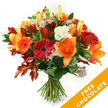 A hand-tied autumn bouquet captured against a clean white studio background, radiating warm, seasonal colour and handcrafted balance. The composition is led by several large, open orange lilies with clear, sculptural trumpets and visible stamens, their petals a vivid, sunlit orange that immediately draw the eye. Surrounding the lilies are plump Gerbera daisies in deep ruby-red and bright orange, their concentric layers of petals forming joyful, graphic discs. Creamy white Lisianthus with softly ruffled edges are tucked in among the bolder blooms, adding a cool, textural contrast that lightens the palette. Small, star-shaped Astrantia with purple-pink centres peek through like delicate pockets of intricate lace, introducing a fine, whimsical texture. Deep green foliage-likely ruscus or salal-forms a lush base, its glossy leaves catching the light and grounding the arrangement. Scattered through the bouquet are striking, deeply lobed oak leaves in burnt orange and reddish-brown, their jagged silhouettes reinforcing the autumn theme and giving the design a natural, gathered-from-the-park feeling. The stems are neatly gathered and tied with dark twine or raffia in a traditional hand-tied style, the knot visible at the base. A bright orange diagonal banner overlays the lower right corner of the image, clearly stating FREE CHOCOLATE in bold white letters, a visual signal of an added seasonal offer. The overall effect is warm, tactile, and celebratory-ideal for sending to a friend on a crisp Stanmore morning or leaving as a vibrant centrepiece on Stanmore High Street, meant to evoke the scent of late-summer fields and the quiet comfort of autumn light.