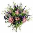 The photograph captures a sumptuous hand-tied bouquet set against a seamless white background, the flowers glowing softly as if illuminated by gentle daylight streaming into a Stanmore bay window. The bouquet is rounded and full, brimming with tones of pink, mauve, purple, and fresh green. At the heart of the design, pastel pink roses open in swirling layers, some fully unfurled with petals drifting from richer blush centres to paler, almost cream edges, others still held in tighter buds that lend the arrangement a sense of life and unfolding stories. Nestled among these are lavish carnations in dusty rose and soft mauve, their finely ruffled petals adding depth, texture, and an almost velvety presence. Punctuating the arrangement, tall spires of deep violet and indigo Veronica stand gracefully, providing height and contrast, a little reminiscent of the elegant vertical lines you might notice walking up Stanmore Hill. Dotted between the larger blooms, clusters of tiny, daisy-like asters in lavender and periwinkle create a light, airy veil, each blossom tipped with a modest yellow centre that brightens the overall look. Rich purple lisianthus flowers deepen the palette, some bells fully open and others just beginning to unfurl, adding a sophisticated edge. Around the blooms, a lush collar of foliage-cool eucalyptus leaves with their silvery sheen, vivid lime-green textural filler that recalls sedum or green trick carnations, and various leafy greens-frames the flowers and infuses the arrangement with freshness. Neatly gathered stems are visible at the base, confirming the hand-tied style and suggesting ease of placing the bouquet in a favourite vase at home in Stanmore. The overall impression is one of gentle opulence and heartfelt intention, perfect for imagining a same-day flower delivery to celebrate a birthday, mark an anniversary, or simply bring comfort and beauty into a quiet evening.
