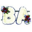 A visually striking and deeply respectful floral tribute forms the capital letters BA, set side-by-side on a low, dark green rectangular base against a clean white background. Each letter is built from hundreds of small, pristine white chrysanthemum florets pressed close together to create a soft, pillowy surface that reads as both delicate and durable. The edges of the B and A are neatly trimmed and framed with a ruffled royal blue satin ribbon, which catches light and gives each letter a crisp, defined silhouette while emphasising the contrast between the pure white face and the vibrant border.

Accents are placed to balance the composition: a lush cluster of velvety deep red roses adorns the upper left curve of the B, and a matching cluster sits at the lower right of the A. Interspersed among the roses are vivid, spike-like blue delphinium-style blooms and small sprays of lighter green trailing foliage that soften the geometry and add a whisper of movement. Darker green leaves tuck behind the blossoms to provide depth and a natural frame. Under soft studio light the white chrysanthemums glow quietly and the roses release a faint, classic perfume, lending the piece a calm, contemplative mood. Crafted with the measured restraint used for memorial work, this pair of initial letters is tailored for a funeral or commemorative service, suitable for display at local chapels, community halls, or quiet corners near Stanmore Common, where families seek a personalised and dignified way to honour a loved one.