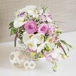 A sophisticated wedding-style floral arrangement in shades of blush pink and pure white is beautifully presented in a round white wicker basket, as if set for a special event in Stanmore. The image shows a close-up view of the arrangement on a crisp white surface, allowing every detail of the flowers to be appreciated. At the heart of the display, large pastel pink roses open in classic spirals, their petals layered and velvety, suggesting softness and gentle fragrance. They are accompanied by white Phalaenopsis orchids, whose broad, satin-like petals extend outward, framing the roses and drawing the eye to their golden-yellow and pale apricot centers, which are touched with the lightest trace of mauve. White Lisianthus blooms, some tinged delicately at the edges with lavender, add a fine, ruffled texture, giving the bouquet an airy, almost cloud-like appearance that would look perfectly at home at a wedding reception near Stanmore Hill or a celebration meal on The Broadway. Throughout the arrangement, dainty white baby's breath blossoms are woven in, their tiny flowers softening the transitions between the larger focal blooms and providing a veil of lightness and detail. Gentle stalks of pale pink stock emerge above the other flowers and then drift slightly over the basket's edge, introducing height and graceful movement, like a subtle cascade across a beautifully laid table. Fresh green foliage, from slender leaves to tiny buds, punctuates the design and introduces vivid contrast, reinforcing the freshly-cut, garden-inspired character. Beside the basket, two decorative spheres woven from pale, natural fibres rest on the white surface, mirroring the basket's texture and adding an understated decorative accent. Behind everything, a neutral beige or taupe fabric background provides a soft, textured setting that allows the colours and shapes of the flowers to stand out. The overall scene conveys refined joy, intimacy, and celebration, ideal for weddings, anniversaries, and elegant gatherings in Stanmore.