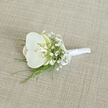 This image shows a refined white orchid boutonnière, carefully arranged as though ready for a groom dressing for a Stanmore wedding near Bentley Priory or along The Broadway. At the heart of the design is a single, immaculate orchid bloom, its rounded white petals smooth and gently curved like folded silk. The centre of the orchid reveals a suggestion of creamy yellow within the throat, adding subtle depth and warmth that softens the pure white. Encircling this focal flower is an airy constellation of white baby's breath, each tiny blossom suspended on fine stems so that the whole cluster looks like a delicate mist or lace overlay. Slender strands of bright green foliage, perhaps fine grass or fern-like leaves, peek out from beneath the bloom, introducing a fresh, linear contrast that frames the softer shapes. All stems are tightly gathered and wrapped in gleaming white satin ribbon, the texture smooth and slightly lustrous, providing a comfortable handle for pinning. Just above the midpoint, the ribbon is tied into a precise, small bow, which adds a note of traditional charm and careful craftsmanship. The boutonnière lies on a light beige, linen-textured fabric that runs diagonally across the image, its fine weave catching light in a way that suggests a quality tablecloth or sample of suiting. The gentle, even lighting casts no harsh shadows, instead bathing the arrangement in a calm glow that conveys serenity, romance, and readiness for a special occasion. It feels like the sort of handcrafted piece a local Stanmore florist would deliver on the morning of a ceremony, quietly waiting to complete a perfectly tailored suit.