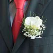 In this image, a wedding boutonnière transforms a classic charcoal suit into something deeply personal and romantic, ideal for a celebration in Stanmore. The design is anchored by a single white orchid resting just above the pocket, its petals broad and velvety, arranged in an open, welcoming shape that draws the eye. Soft light washes over the bloom, catching on its creamy surface and revealing a delicate heart tinted with gentle gold and blush hues, like a whisper of warmth at the centre of calm. Surrounding the orchid is a cloud of baby's breath, tiny white flowers clustered in an airy ring that softens the overall silhouette and adds a sense of ethereal lightness. Interspersed among them, slim stems of greenery and wispy fern fronds bring in refreshing green tones, reminiscent of the parks and quiet, leafy corners around Stanmore. The flowers are expertly gathered and secured at the base with a narrow white ribbon, folded smoothly and tied like a miniature ceremonial sash, showing the care taken in every detail. The boutonnière sits neatly within the pocket of a dark, tailored jacket, whose clean lines and rich texture provide a sophisticated backdrop. A striking red tie runs down the centre, adding vibrancy and a contemporary edge that hints at joy, passion, and celebration. You can almost imagine the wearer adjusting the lapel in a mirror before heading to a Stanmore venue, perhaps a local church or reception hall, ready for photographs and heartfelt vows. This floral piece feels like a quiet promise pinned close to the heart, symbolising love, hope, and the beauty of a day shared with family and friends.