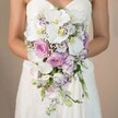 A radiant bride from Stanmore, her arms gently curved and gown adorned in classic lace, tenderly offers a magnificent cascading bridal bouquet, set against a backdrop of creamy beige reminiscent of soft dawn light near Stanmore station. The bouquet's generous teardrop shape brims with the gentle hues of white, powder pink, and subtle lavender-each flower carefully chosen to embody the refined romance that characterizes Stanmore weddings. Majestic phalaenopsis orchids, their elegant white petals softly arching and yellow centers aglow, take center stage, surrounded by plush pink garden roses and airy lavender lisianthus. Wispy sprigs of baby's breath, with their feathery whiteness, are scattered among the blooms, mingling with delicate green foliage reminiscent of garden walks through Bernays Gardens. The bridal gown's intricate bodice peeks from beneath, hinting at craftsmanship and tradition cherished in local ceremonies. This expertly arranged bouquet, lovingly assembled by Stanmore's finest florists and available for delivery, radiates sophistication and emotional warmth-a contemporary yet classic floral statement for North London's most treasured wedding celebrations.