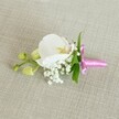 A delicately crafted boutonniere is laid horizontally on a softly textured, oatmeal-coloured linen background, the neutral fabric giving the whole scene a calm, refined air reminiscent of a quiet morning in Stanmore before a wedding at a local church or registry office. At the heart of the design sits a single, pristine white Phalaenopsis orchid, its broad, velvety petals opening in a graceful curve. The orchid's centre glows with subtle tones of soft yellow and the faintest blush of pale pink, drawing the eye to its intricate throat. Around the base of this central bloom, tiny clusters of white baby's breath form a cloud-like halo, their minuscule blossoms adding a gentle, airy texture that feels almost weightless against the linen. Slender, vibrant green leaves peek out from behind, offering a fresh contrast to the white petals and echoing the greenery of local Stanmore gardens and nearby parks. To one side, a short stem carries a couple of tightly closed, lime-green orchid buds, suggesting the quiet promise of blooms yet to open, like a new chapter at a prom or engagement celebration. All the stems are neatly bound together with a slim, bright fuchsia pink satin ribbon, tied into a small, elegant bow whose glossy sheen catches the soft, even light. The lighting is gentle and diffused, highlighting the smooth orchid petals, the delicate spheres of the Gypsophila, and the subtle weave of the linen. This refined floral accessory feels ideal for pinning to a groom's lapel, a groomsman's jacket, or a special guest's outfit for a formal event in and around Stanmore.