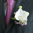 A close, intimate view of a groom's charcoal suit lapel in Stanmore shows a meticulously crafted white Phalaenopsis orchid boutonniere glowing against the dark fabric. The suit jacket is sharply tailored in deep charcoal, almost black, with a finely woven texture that catches the light in soft, controlled highlights. Just beneath the lapel, a smooth mauve tie in rich magenta-plum tones is neatly knotted over a pale grey dress shirt with a faint, textured pattern, giving the whole look a sense of quiet luxury suited to weddings in local Stanmore venues. Pinned to the left lapel, the orchid boutonniere is the undeniable focal point: broad, pristine white petals curve gently outward, satiny and slightly undulating like silk in a light breeze, framing a golden-yellow and soft orange centre that adds warmth to the composition. Around the orchid, dainty sprigs of pale lilac blossoms and clusters of tiny white gypsophila-baby's breath-create a misty halo of texture, suggesting a meadow distilled into miniature. Fresh, deep green leaves cradle the flowers and give structure to the arrangement, with a single pale green orchid bud standing upright above the main bloom, hinting at new beginnings. At the base, a slim lavender satin ribbon is tied into a small, perfectly formed bow, its sheen echoing the tie's hue and drawing the eye toward the suit's breast pocket. The lighting is soft and flattering, evoking the calm anticipation before a ceremony, like a groom stepping out near Stanmore Hill or Bentley Priory for those first photographs. This image conveys understated elegance and romance, a small yet powerful floral detail that feels both personal and timeless, as if chosen with great care from a trusted local florist on Church Road.