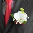 A close-up photograph focuses on a single wedding boutonniere pinned neatly to the lapel of a dark charcoal suit, the fine woven texture of the jacket clearly visible beneath the flowers. At the centre, a large white Phalaenopsis orchid opens its wide, velvety petals, the creamy white subtly catching the light, while the orchid's yellow-and-white lip provides a soft, golden focal point. Nestled just above, two small, round lime-green chrysanthemum-style blooms add a fresh, zesty contrast, their compact petals forming tight domes of colour. Around the orchid, delicate sprigs of white baby's breath scatter tiny cloud-like blossoms, softening the edges of the design and giving it a romantic, bridal feel. Rich green leaves cradle the flowers, adding depth and a natural frame against the dark wool suit. To the left of the boutonniere, a solid red satin tie stands out boldly over a light grey and white checked dress shirt, the tie's sheen echoing the fine deep red ribbon wrapped around the stem of the boutonniere. The flowers sit just above a precisely stitched breast pocket, suggesting a carefully tailored outfit ready for a wedding ceremony, perhaps at a Stanmore synagogue or a venue near Stanmore Country Park. The lighting is gentle and flattering, revealing each curve of the orchid and the subtle gloss of the foliage. The overall mood is refined and intimate, as if the wearer is taking a quiet moment before walking down the aisle, the floral detail capturing love, commitment, and the polished craftsmanship you'd expect from an experienced Stanmore wedding florist.