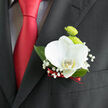 This image focuses on a single, carefully crafted boutonnière resting against the lapel of a dark, tailored suit, ideal for a sophisticated Stanmore celebration. At its centre, a white orchid blooms with serene, sculptural beauty, each petal wide, smooth and gently curved, giving the impression of fine porcelain warmed by soft daylight. Its core holds a touch of pale yellow and light green, offering a subtle glow that draws the eye inward. Just behind the orchid, two lime-green button chrysanthemums rise like textured spheres, their many tiny petals forming dense, rounded domes that add youthful vibrancy and a modern twist. Encircling the flowers, slender sprays of baby's breath carry dozens of miniature white blossoms, creating a delicate, airy halo that softens the transitions between flower, foliage and fabric. Deep green leaves cradle this cluster, their polished surfaces and clean lines giving structure and ensuring the colours remain crisp against the charcoal-black suit. At the base of the stems, a slim ribbon in rich burgundy-red is expertly wrapped, providing a refined finishing touch that mirrors the deep red satin of the tie below. The whole arrangement exudes quiet confidence, as if it has been prepared by an experienced local florist in Stanmore who understands how flowers must endure a full day of festivities-from a heartfelt ceremony on Marsh Lane to relaxed photos in Canons Park. The boutonnière feels personal and intentional, the kind of floral detail chosen to express character and care without overshadowing the overall look, adding a final note of romance to the wearer's ensemble.