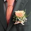 Pinned to the left lapel of a deep charcoal suit jacket, a single peach-pink rose commands attention with gentle modesty, its petals unfolding in layers that catch the soft daylight as if lit from within. Surrounding the rose, delicate sprigs of white baby's breath (gypsophila) float like tiny clouds, while slender, airy green foliage threads through the arrangement to add movement and texture. The boutonnière's base is finished with a slim, lustrous peach satin ribbon tied with careful restraint, harmonising with the similarly hued tie that lies flat over a pale dress shirt patterned with a subtle dark grid. Texturally, the matte wool of the suit, the satiny sheen of the ribbon, and the velvety surface of the bloom form a quiet trio that reads as both classic and handcrafted. The visual impression suggests a soft, slightly sweet scent - an ephemeral whisper rather than a heavy perfume - and an intimacy suited to weddings, civil partnerships, or formal family celebrations. As a Stanmore florist I often assemble variations like this for men preparing to walk down Stanmore High Street or to stand beside loved ones at local venues; this particular composition reflects that local sensibility: modest, well-coordinated, and seasonally fresh. Prepared with attention to proportion and pace, the boutonnière carries the same careful restraint I use in my shop, Flowers Stanmore, where textures and colour are chosen to flatter both the wearer and the setting without stealing the moment.