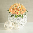 A hand-arranged sphere of peach roses and soft gypsophila sits upon a white woven orb, an object that reads as both floral and sculptural when I prepare it at Flowers Stanmore near Stanmore Broadway. The roses gather in a low, luxurious dome, petals folding into buttery spirals that shift from pale cream to honeyed apricot toward the edges, each bloom suggesting tenderness and quiet celebration. Clouds of tiny white baby's breath lace between the roses, blurring edges and giving the bouquet an ethereal, veil-like softness that sparkles where light meets blossom. Rich green foliage frames the composition, lending contrast and the faint green scent of freshly cut leaves beneath the sweeter perfume of roses. The vessel is a crisp white rattan sphere with an open lattice; its texture and negative space make the arrangement seem light and contemporary, while two small, airy spheres placed beside it create a rhythmic visual echo. Late morning light, like that which filters through windows on Elm Park Road or drifts across the platforms at Stanmore Station, warms the petals and brings a subtle golden glow to the scene. This piece suits intimate weddings, milestone anniversaries, or a serene home centrepiece, offering a sense of calm and new beginnings. Thoughtfully composed for local delivery across Stanmore and nearby Canons Park, it reflects the care and craft of an experienced local florist and the particular softness of suburban London light.