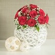 A refined table arrangement dominates this image, centred on a rounded bouquet of deep, velvety red roses that appear to glow softly against a pale backdrop. The roses are packed closely together, forming a smooth dome where each bloom opens fully, petals layered in tight spirals that create a sense of depth and gentle movement. Woven delicately among the crimson flowers are airy sprays of white baby's breath, their tiny blossoms scattered like stars against a night sky, lightening the overall palette and adding a romantic, almost ethereal quality. Small accents of green foliage peek out from between the flowers, offering subtle contrast and reminding the viewer of the arrangement's natural origins. The bouquet is gathered into a distinctive round vase constructed from curved white strands that twist and cross in an open-weave design, much like a modern white wicker sphere. This sculptural container provides texture and structure, cradling the flowers while allowing glimpses of the stems within. Resting to the left on the smooth white surface are two decorative spheres in soft cream tones, woven from flat strips into open lattices, the larger sphere placed nearer the viewer to enhance the sense of depth and harmony. The background is a softly blurred, neutral beige wall with a fine, fabric-like pattern that stays gently in the distance, ensuring the vivid reds and clean whites remain the visual focus. The overall impression is of a thoughtfully styled centrepiece, ideal for a Stanmore wedding table, a romantic dinner near Bentley Priory, or an elegant home display, showing the kind of refined composition that local florists in Stanmore create for special occasions.