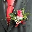 Detailed close-up of a romantic wedding boutonniere resting against a charcoal grey suit jacket, evoking the polished calm of a Stanmore wedding morning. The heart of the arrangement is a single cerise-pink rosebud, partially opened so its velvety petals curl inward in a refined spiral, softly catching the light against the darker wool. Surrounding the rose are clusters of white baby's breath, their diminutive blooms forming a gentle, airy cloud that contrasts with the bold central hue and lends an almost ethereal quality. Slender, feather-like sprigs of greenery reach outward from behind the flowers, arranged alongside broader, glossy green leaves that create a balanced, layered frame, reminiscent of the lush borders found in gardens around Stanmore. At the base of the boutonniere, a bright scarlet satin ribbon is tied into a small, elegant bow, its subtle shine mirroring the smooth red silk tie visible under the lapel. The tie lies against a grey dress shirt with a faint, sophisticated pattern, adding depth without drawing focus away from the floral detail. The suit's fine wool blend appears slightly textured, offering a tactile counterpoint to the softness of the petals and the delicate structure of the gypsophila. This buttonhole feels tailored for meaningful vows at a Stanmore church, synagogue, or registry office, capturing a blend of tradition and modern elegance. It suggests a wearer standing with quiet confidence, greeting guests and stepping into photographs, the flowers serving as a small but powerful emblem of love, hope, and the shared journey that begins on this special day.