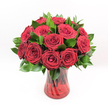A luxurious bouquet of deep crimson red roses stands at the centre of the image, every bloom opened to a generous, velvety swirl of petals that spiral from a tight heart to softly unfurling outer edges. The roses are densely clustered, forming a rounded, opulent dome of colour, with no gaps between the flowers, just a sea of rich ruby tones that immediately suggests romance and devotion. Around and between the roses, glossy dark green foliage, likely Ruscus or a similar leaf, adds depth and structure, its smooth surfaces catching the light like polished satin and giving a fresh contrast to the plush petals. The entire arrangement is cradled in a translucent ruby-red glass vase, its sides subtly twisted into a spiralled design that ripples with reflections. The glass is rich but not opaque, allowing the green rose stems to be seen through the red tint, creating layers of warm colour from base to bloom. The vase widens slightly at the top, providing a stable, elegant shape that grounds the dramatic flowers. Bright, even lighting falls from above, causing each petal edge and leaf vein to stand out clearly, while the pure white background removes all distraction and makes the bouquet the sole point of focus. The impression is of a classic romantic gift, the sort of arrangement you might send in Stanmore for a Valentine's surprise, a milestone anniversary on Church Road, or a heartfelt declaration of love shared quietly at home. Though the photograph is silent, you can almost imagine the faint, sweet fragrance of the roses, wrapping the moment in tenderness and sincere emotion.