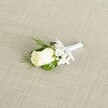 A classic white rose boutonniere lies diagonally across a soft, linen-like beige fabric, its refined simplicity echoing the quiet elegance of a Stanmore wedding. At the heart of the design is a single creamy white rosebud, petals still gently furled, their velvety texture catching the soft, diffused light. The bloom feels fresh and just-opened, with a hint of pale green at the base where the sepals cup the flower, suggesting it has been prepared only moments ago by a careful florist. Around the rose, tiny clouds of white baby's breath float delicately, each miniature blossom adding lightness and a whisper of texture, like fine lace against the lapel of a well-cut suit. Slender, deep green leaves arc gracefully behind the flowers, some glossy and smooth, others more feathery, giving the boutonniere depth and a natural frame. The stems are neatly bound in a pristine white satin ribbon, wrapped tightly and finished with a small, tidy bow that sits just below the bloom, ready to be pinned in place for a ceremony at a Stanmore synagogue, church, or registry office. The neutral, finely woven fabric beneath has a subtle diagonal pattern, resembling raw silk or lightly textured linen, offering a warm, understated backdrop that allows the white rose to glow. Gentle, even lighting softens any shadow, emphasising the purity of the petals and the calm, hopeful mood of the piece. This image captures the intimate detail of groom or groomsmen flowers, as one might see them arranged for a local celebration near Stanmore Station or along The Broadway, symbolising new beginnings, promise, and timeless devotion.