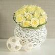 The photograph captures a serene and stylish floral centerpiece, the sort of design a local Stanmore florist might prepare for a wedding breakfast or intimate celebration near Belmont Circle. At the top of the arrangement, a dome of light yellow roses forms a smooth, rounded mass. Each rose is open and full, revealing ruffled layers of petals that curve outward in soft spirals, creating a cushion of velvety texture. The colour rests between buttery cream and pale lemon, bathing the bouquet in a gentle, warm light. Nestled among these roses are fine sprigs of white baby's breath, their many tiny blossoms perched on slender stems, giving the bouquet a delicate, cloud-like veil and adding subtle depth. Encircling the base of the floral dome, a tidy band of fresh green foliage wraps around like a slim leafy belt, clearly marking the transition from blooms to container and offering lively contrast. The arrangement is set in a white woven vase, crafted from thin strands that cross over one another in a loose, open pattern. The vase broadens at the top and bottom while narrowing in the middle, its silhouette echoing the soft curves of the roses above. The open weave allows soft shadows to fall across the smooth, reflective white table surface, where two pale decorative spheres lie to one side. These orbs are made from similarly interlaced strips, forming airy, geometric shapes that complement the vase's texture. The background fades into a gently blurred wash of warm beige, suggesting a quiet, well-kept interior without pulling attention from the focal arrangement. Together, the flowers, vase, and decorative spheres create a mood of elegant simplicity and subtle romance, perfectly suited to a Stanmore dinner table or wedding reception styled with understated charm.