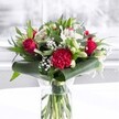A rich and romantic bouquet sits proudly in a clear cylindrical glass vase, the freshly cut green stems visible through the water like a neat bundle of ribbons, suggesting great care from the florist's hand. Deep red roses, their velvety petals just beginning to unfurl, are clustered at the heart of the arrangement, radiating passion and timeless elegance. Beside them, a large, ruffled crimson carnation anchors the front, its many frilled layers catching the soft light and adding a dramatic, almost tactile texture. Scattered between these bold reds are creamy white blooms: delicate white alstroemeria with fine speckles in their throats, petite white chrysanthemums, and closed buds of lilies that hint at future blossoms. Wisps of baby's breath float like tiny stars throughout, giving the whole bouquet an ethereal softness and a sense of generous fullness. Around the base, broad, glossy dark green leaves are wrapped and gently curled to form a sophisticated collar, while finer eucalyptus-style foliage and slender pointed leaves weave upwards, bringing movement and depth in several fresh shades of green. The arrangement is photographed on a pristine white surface, with a softly blurred, bright background that feels like daylight streaming through a Stanmore window, perhaps looking out towards Bentley Priory or a quiet residential street off The Broadway. The lighting is gentle and even, allowing each petal edge and leaf vein to be seen clearly, creating an atmosphere of refined celebration. It feels like a bouquet chosen to grace an anniversary dinner, a milestone birthday, or a thoughtful delivery to a Stanmore home or office, offering warmth, gratitude, and heartfelt affection in one carefully balanced display of red, white, and green.