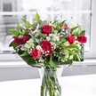 A lush bouquet of rich red roses, ruffled carnations, white alstroemeria, and clouds of baby's breath stands in a clear glass vase on a bright white tabletop, lit by soft daylight from a large window. The roses are a deep crimson, some fully open to reveal velvety, spiralled petals, others still in elegant bud form that promise to unfurl in the coming days. Nestled closely among them, the carnations share the same bold red tone, their frilled petals creating a sense of fullness and traditional romance. Between the warmer blooms, white alstroemeria stems rise with a mix of tight buds and partially opened flowers, their trumpet-shaped petals tipped with pale green that echoes the fresh foliage below. Tiny white gypsophila blossoms float through the design like a delicate mist, softening the transitions between the stronger colours. The bouquet is cradled by broad, glossy green leaves at its base, giving the arrangement a sculpted, polished outline, while slimmer stems of leafy greenery weave through the centre, adding movement and depth. Through the cylindrical glass vase, you can see the neatly trimmed stems fanning out in clear water, reinforcing the sense of freshness as if the flowers were just conditioned in a Stanmore workshop before delivery. The white tabletop reflects a faint halo of light back up into the heart of the arrangement, while the outdoor scene beyond the window is blurred and almost monochrome, making the reds, whites, and greens appear even more vivid. The overall feeling is of a joyful, heartfelt gift-perfect for birthdays, anniversaries, or simply brightening a home near Stanmore station on a clear morning.