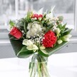 A contemporary mixed bouquet glows in a clear glass vase, positioned near a softly lit window that could easily be overlooking the quiet streets of Stanmore on a bright morning. At the heart of the arrangement, a velvety coral-red rose and a richly ruffled crimson carnation provide a dramatic focal point, their deep tones pulsing with warmth and emotion. Around them, clusters of delicate white chrysanthemums, daisy-like blooms, and tiny button flowers create a halo of lightness, their petals crisp and feathery, like freshly opened blossoms in spring. Creamy white lilies nestle between the smaller blooms, some fully open with star-shaped petals, others still in elegant bud, promising more beauty to come over the next few days. Broad, glossy emerald leaves sweep in graceful arcs around the base, expertly folded and layered by a skilled florist to cradle the flowers and give the design a clean, modern silhouette. The glass vase reveals the neat stems beneath the waterline, reinforcing the sense of freshness, as though this bouquet has just arrived from a local Stanmore flower market. Gentle daylight washes over the arrangement, catching the sheen of the foliage and the subtle gradations of red and white, and giving the whole display an airy, uplifting presence. You can almost imagine the mingled scent: a blend of sweet lily, softly spiced carnation, and leafy green notes that would quietly fill a living room or hallway. This is the kind of bouquet you might place on a dining table for a family celebration in Stanmore, or send as a thoughtful, cheering gift to brighten an ordinary weekday for someone you care about.