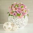 Evoking the tranquil elegance of Stanmore's leafy lanes and garden celebrations, this handcrafted floral centerpiece graces a white table under soft natural lighting. The arrangement springs from an artistically woven white vase, its open, lattice-like structure conveying both contemporary flair and rustic charm, reminiscent of the local craft found at Stanmore Village fairs. Within, roses in shades of creamy pink and soothing lavender unfurl their plush petals, complemented by demure pink alstroemeria showing off intricate, softly striped throats. Clumps of lively lime green button chrysanthemums infuse a refreshing touch amidst sprays of delicate baby's breath, which dance like morning mist over Canons Drive. Deep green foliage tucks between the blooms, anchoring them visually and emotionally in the lush spirit of Stanmore's parklands. Resting alongside, two decorative spheres woven from pale, organic tines echo the basketry, their interplay of sizes and textures adding visual balance and a whimsical, handmade feel. The beige, subtly textured backdrop blurs softly behind the bouquet, placing all focus on the artistry and abundance at the foreground. Together, they form not just a wedding table arrangement, but a celebration of place and skill-timeless, heartwarming, and unmistakably crafted for Stanmore's most cherished gatherings.