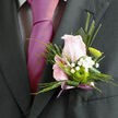 The photo presents an artfully composed boutonnière, the kind of detail a Stanmore groom might choose to express quiet elegance on his wedding day. At its heart is a single pale pink rose, just beginning to unfurl, with velvety petals layered in soft spirals that convey warmth and tenderness without being overly showy. Encircling the rose are delicate sprays of white gypsophila, their tiny blossoms scattered like starlight, adding a dreamy, cloud-like softness and a touch of innocence to the overall design. A neatly formed green chrysanthemum button sits close to the rose, its vivid lime hue and compact, textured petals bringing a fresh, contemporary twist that subtly updates the classic rose-and-gypsophila pairing. Slender, architectural pieces of greenery and a carefully folded dark leaf create a clean, tapered line along the stem, making the boutonnière feel tailored and refined-perfect for a well-fitted suit worn at a Stanmore registry office or a nearby country house venue. The stems are bound together in a soft mauve satin ribbon, wrapped smoothly with a slight, graceful tail, the muted lavender tone echoing the romantic notes of the central flower. The interplay of colours-blush, white, green, and gentle lilac-feels perfectly balanced, neither too bold nor too pale. Imagining it pinned to a lapel near Stanmore station or at a ceremony overlooking local parks, you can sense the quiet confidence it lends, the faint floral scent that calms pre-ceremony nerves, and the way this small, handcrafted piece of floral art speaks gently of care, commitment, and thoughtful preparation.