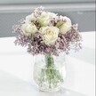 A softly lit close-up of a serene floral arrangement shows creamy white roses opening gently above a cloud of lavender-purple statice, captured as if sitting on a bright windowsill in a quiet Stanmore home. Each rose is full and fresh, its velvety petals unfolding in loose spirals, with a faint hint of pale yellow at the very centre that suggests it has only just come into perfect bloom. Around them, tiny papery blossoms of statice form a delicate halo of misty lilac, their branching stems creating a feathery, textured cushion that makes the roses feel both supported and beautifully framed. The bouquet is gathered in a clear glass vase with an ice-crackle texture, so the surface seems to shimmer and fracture the light, like thin frost on a winter morning on Stanmore Hill. The water inside is clean and transparent, revealing straight green stems that cross and angle gracefully, adding a fresh, botanical rhythm to the design. The vase rests on a smooth, white surface that reflects a faint echo of the arrangement, casting pale, soft-edged shadows that enhance the overall sense of calm. Behind, the background fades into a blurred wash of light grey, completely unobtrusive, so the eye remains on the gentle interplay of white, lilac, and green. The mood is one of quiet comfort and understated elegance, well suited to a thoughtful get-well message, a peaceful condolence bouquet, or a simple gesture of affection delivered to a cosy flat near Stanmore station or a calm office overlooking Canons Park. Every element in the image emphasises simplicity, purity, and the soothing presence of fresh flowers indoors.