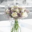 A rounded bouquet of creamy white roses and soft lavender statice stands calmly at the centre of the image, its shape full and perfectly balanced, like a gentle cloud of petals captured in a quiet moment. Around a dozen white roses, their velvety blooms just opening, reveal spirals of ivory-toned petals that catch the light with a subtle sheen. Threaded between them, countless tiny lilac-purple flowers, likely statice, create a fine, misty halo, their papery texture adding delicate contrast to the plush depth of each rose. The flowers are gathered in a tall, clear cylindrical glass vase, the stems straight and fresh, visible through the water where pale green hues deepen towards the base. The vase is placed on a spotless white surface that softly reflects the arrangement, suggesting a tidy console table or countertop in a modern Stanmore home or office close to Canons Park. Diffused daylight streams from an unseen window, bathing the bouquet in a cool, even glow that highlights the creamy whites and gentle purple tones without any harsh shadows. The overall impression is serene and composed, ideal for a peaceful sympathy gesture, a quiet anniversary surprise, or a calming presence in a waiting room near Stanmore Hill. The bouquet feels freshly arranged, as though a local florist has just set it down after a final check of every stem, ensuring that the roses face outward and the statice surrounds them like a soft, protective aura. In this still scene, the flowers seem to offer a gentle pause, a moment to breathe, reflect, and feel quietly comforted by natural beauty.