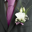 This photograph captures a refined wedding boutonnière pinned neatly to a dark, structured suit, an understated yet expressive detail perfect for a Stanmore ceremony. At the centre, a pristine white lisianthus flower unfolds in layered, slightly ruffled petals that spiral gently outward, creating a soft focus point against the charcoal fabric. The texture of the petals looks almost like fine tissue or silk, inviting the imagination to feel their cool, velvety surface. Behind the main bloom, two lisianthus buds stand gracefully, still wrapped in their green sepals, their elongated shapes introducing a sense of height and quiet anticipation. Around the central flower, delicate sprigs of baby's breath create a light, misty framework, their countless miniature blossoms dotting the boutonnière with flecks of white that resemble tiny stars. Wisps of narrow green foliage thread through the arrangement, some curving gently, others more straight and architectural, giving the piece a balanced mix of formality and natural, garden-inspired ease. At the base, a single glossy leaf is rolled into a smooth curve, cradling the stems and bringing visual stability, much like the sturdy greenery you might see in well-kept Stanmore front gardens. The stems themselves are wrapped snugly in a soft mauve ribbon, secured in a neat bow whose colour subtly nods to a lavender or lilac tie worn by the groom. The overall impression is of clean, thoughtful elegance and a faint, fresh floral scent-an accent that feels personal and timeless. It's the kind of boutonnière that suits both a traditional church wedding in Stanmore and a more intimate local gathering, carrying with it a sense of joy and new beginnings.