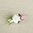 A delicately crafted wedding boutonnière rests at a gentle diagonal on a softly textured, light beige fabric that looks like finely woven linen, its warm neutral tones echoing the calm of a Stanmore morning. At the heart of the design is a single white lisianthus flower, fully open and velvety, its ruffled petals forming a rounded, cloud-like bloom that feels both romantic and refined. Just beside it sit two pale cream and green lisianthus buds, still tightly closed and pointing slightly downward, suggesting quiet anticipation and the promise of a new chapter. Tiny sprays of white gypsophila are threaded between the blooms, their miniature starry flowers creating a soft halo and adding a whisper-light fullness that catches the eye without overwhelming the central blossom. Slender, grass-like blades of fresh green foliage sweep gracefully to the left, stretching beyond the flowers as if stirred by a gentle breeze blowing across Stanmore Common, while a broader, darker green leaf curves behind the lisianthus, giving the arrangement a sense of structure and depth. The stems are neatly gathered and bound at the lower right with a vibrant fuchsia satin ribbon, tied into a small and jaunty bow; its silky texture and bold pink colour provide a joyful flash against the muted backdrop. The lighting is soft and even, revealing every petal fold, leaf vein, and weave in the fabric, creating an intimate, serene mood. This lapel-sized floral piece feels perfect for a groom, best man, or stylish prom guest in Stanmore, capturing the essence of a special day in a compact, elegant flourish of white, green, and bright pink.