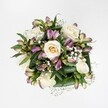 A top-down view reveals a compact, hand-tied bouquet laid against a crisp white background, every petal and leaf standing out clearly as if resting on fresh Stanmore snow. At the heart of the arrangement, creamy white and soft ivory roses unfurl in various stages of bloom, from tight buds with a hint of buttery yellow at their centres to fully opened roses whose layered petals spiral outwards with quiet grace. Between these roses, clusters of mauve and purplish-pink alstroemeria peek through, their slender petals brushed with lavender tones and fine green streaks, like twilight skies over Stanmore Common translated into petal form. Sprays of tiny white gypsophila drift delicately around the focal blooms, creating a cloud-like halo that softens every edge and adds a sense of airiness and romance. A collar of deep, glossy green foliage encircles the flowers, the broad leaves folded and curved to cradle the blooms, giving structure and richness to the overall shape. The lighting is bright yet gentle, catching the velvety texture of the rose petals and the satin sheen of the alstroemeria, suggesting freshly prepared flowers ready for same-day delivery across Stanmore and nearby Canons Park. The mood is serene and elegant, ideal for marking an anniversary, sending thoughtful congratulations, or offering comfort with a refined, modern touch. Every detail in this bouquet reflects the sensitive eye of an experienced florist, balancing colour, texture, and space so the arrangement feels both full and breathable, as if holding a moment of calm in the middle of a busy Stanmore day.